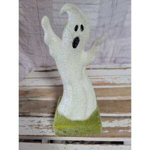 May include: A white glitter ghost with black eyes and a mouth, standing on a green grass base. The ghost is a Halloween decoration.