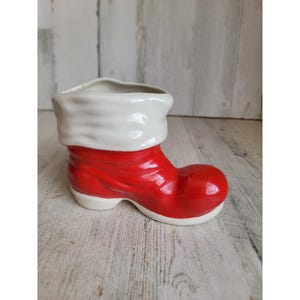 May include: A ceramic boot-shaped planter or container featuring a glossy red lower section and a white cuff at the top. The design mimics a classic boot with a rounded toe and a smooth, reflective finish on the exterior surface of the ceramic.
