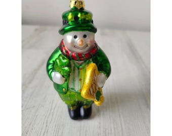 Peut inclure: Une décoration de Noël en verre vert représentant un bonhomme de neige habillé en lutin. Il porte un chapeau vert, une écharpe rouge et tient un trèfle et une harpe dorée.
