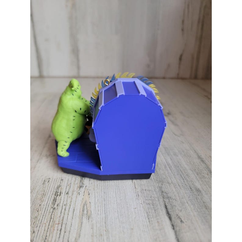May include: A green, stitched, Christmas ornament of the character Oogie Boogie from the movie The Nightmare Before Christmas. The ornament is in the shape of a coffin with a blue and purple exterior. The ornament is decorated with gold glitter.