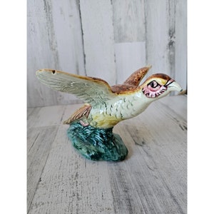May include: A ceramic figurine of a brown and white bird with outstretched wings, perched on a green base. The bird has a pink eye and a brown beak.