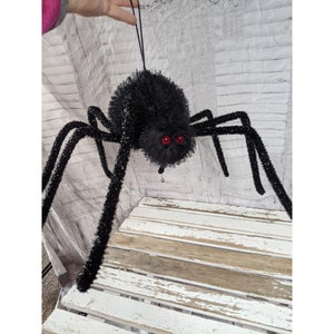 Puede incluir: Una araña negra grande con ojos rojos y patas largas y peludas. La araña está colgada de una cuerda y está hecha de un material suave y peludo.