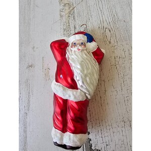 May include: A festive Christmas ornament depicting Santa Claus. The ornament is red with white trim and a long white beard. Santa is wearing a blue hat and carrying a sack. The ornament has a shiny, reflective surface, typical of Christmas decorations.