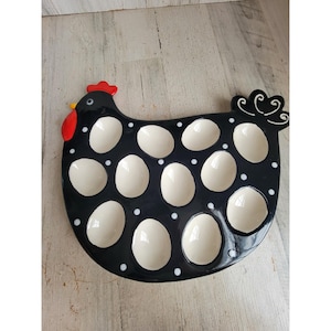 May include: A black ceramic egg tray shaped like a chicken with a red comb and wattle. The tray features twelve cream-colored circular indentations for holding eggs, decorated with small white polka dots across the black surface of the chicken.