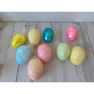 May include: A collection of colorful, glitter-covered Easter eggs. The eggs are in various pastel shades, including yellow, blue, pink, and green. Each egg has a small ribbon loop for hanging. The eggs are arranged on a light-colored wooden surface.