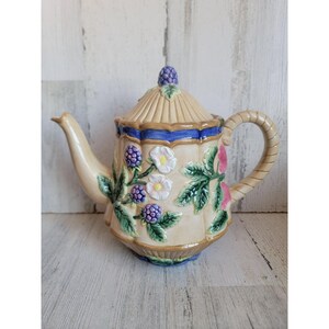 Op de afbeelding: Een theepot van keramiek met een crèmekleurig lichaam en een blauwe en gouden rand. De theepot is versierd met een bloemmotief met roze, witte en paarse bloemen, groene bladeren en bramen.