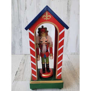May include: A wooden nutcracker figurine in a red and white striped house with a green base. The nutcracker is wearing a red and gold uniform with a white beard and a blue hat.