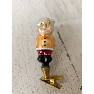 Puede incluir: Un adorno navideño de vidrio vintage con forma de persona que lleva un abrigo amarillo, pantalones rojos y un sombrero verde. El adorno tiene un clip de metal dorado en la parte inferior.