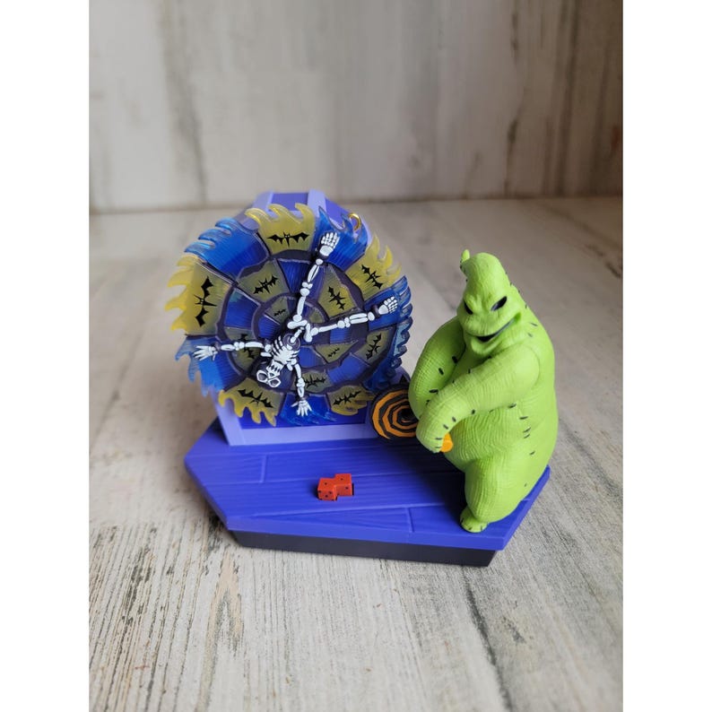 May include: A green, stitched, cartoon character with a toothy grin, holding a spinning wheel with a skeleton on it. The wheel is blue and yellow with black bats. The character is standing on a purple platform with a red brick.