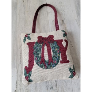 May include: A decorative pillow with the word "JOY" in burgundy letters. The "O" is a green wreath with a red bow. The pillow has a burgundy handle and holly accents. The background is a light beige.