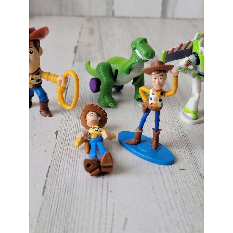 May include: A collection of Toy Story character figurines, including Woody, Buzz Lightyear, and Rex the dinosaur. Woody is featured in multiple poses, with one riding a blue surfboard. The figures are set against a white wooden background.