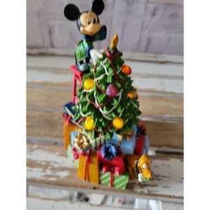 May include: A Christmas tree figurine with Mickey Mouse decorating it. The tree is surrounded by presents and a small dog figurine. The figurine is made of plastic and has a painted finish.