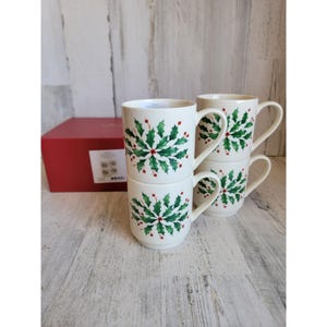Könnte beinhalten: Vier weiße Keramikbecher mit einem grünen Stechpalmen- und roten Beerenmuster. Die Becher sind gestapelt und haben gebogene Griffe. Eine rote Geschenkbox ist zu sehen, was auf ein festliches, saisonales Thema hindeutet.
