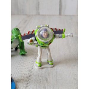 May include: A Buzz Lightyear action figure in a green and white spacesuit with a clear helmet. The figure has its arms outstretched and wings extended. A green dinosaur toy is partially visible in the background.