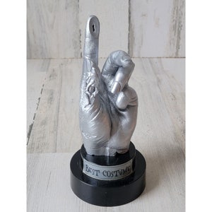 May include: A silver hand sculpture with a raised middle finger, mounted on a black base with the words "Best Costume" inscribed on a silver band.