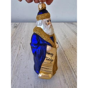 May include: A glass Christmas ornament shaped like a figure in a blue robe with a white beard and a gold-colored collar. The ornament has a gold-colored hat and is holding a scroll that reads "Henry Teddy".