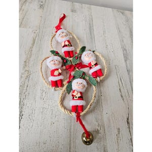 May include: A Christmas decoration featuring four Santa Claus figures hanging from a rope circle with a bell at the bottom. The Santas are wearing red suits with white trim and have white beards. The rope circle is decorated with green leaves and red berries.