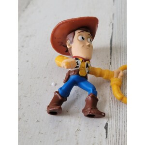 May include: A small plastic figurine of Woody from the Toy Story franchise. The figure is wearing a brown cowboy hat, yellow shirt, blue pants, and brown boots. He is holding a yellow lasso.