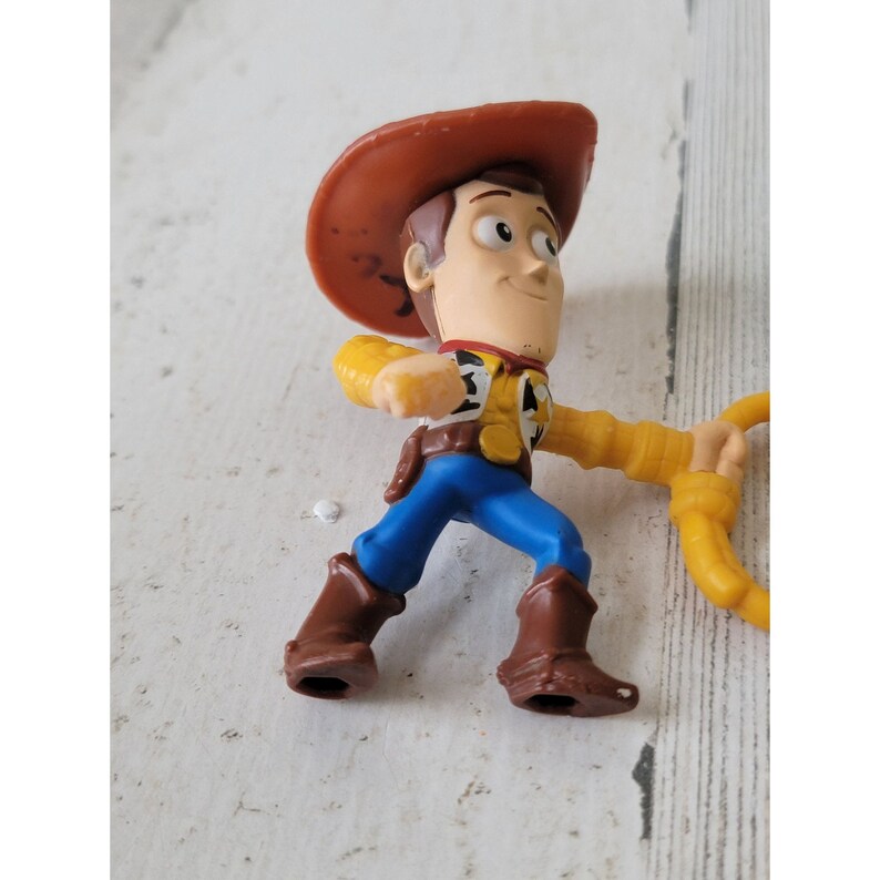 May include: A small plastic figurine of Woody from the Toy Story franchise. The figure is wearing a brown cowboy hat, yellow shirt, blue pants, and brown boots. He is holding a yellow lasso.