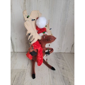 May include: A brown moose with large, cream-colored antlers is wearing a red scarf and a Santa hat with a white pom-pom. The moose is decorated with colorful lights.