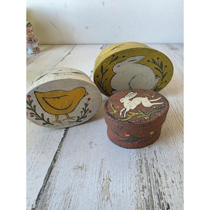 May include: Three decorative, hand-painted ceramic boxes. One is oval with a yellow chick design, another is round with a white rabbit, and the third is a small, round box with a white rabbit. The boxes are painted with earthy tones and botanical accents.