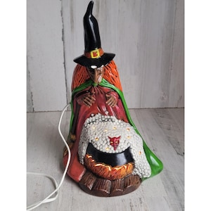 May include: A ceramic witch figurine wearing a black hat and a green and red robe, sitting by a cauldron with a white, fluffy blanket and a red devil face inside. The cauldron is lit with orange flames and the witch is holding a white, fluffy blanket.