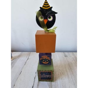 Könnte beinhalten: Ein Halloween-gestuftes Mittelstück mit einer schwarzen Eule, die einen Partyhut trägt, einem lila Würfel mit einem Kürbisgesicht und einem grünen Würfel mit dem Wort "Halloween" darauf.