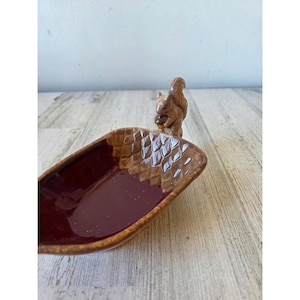 May include: A brown ceramic dish with a squirrel figurine on the end. The dish has a deep burgundy interior and a textured, diamond-patterned rim. The squirrel is holding a nut. The dish is on a light-coloured wooden surface.