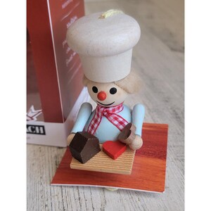 May include: A wooden figurine of a chef wearing a tall white hat, a light blue shirt, and a red and white checkered scarf. The figure holds a small wooden board with a dark brown cube and a red heart-shaped piece, standing on a red base.