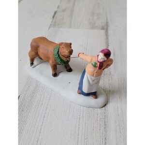 May include: A brown cow figurine wearing a green wreath around its neck is being led by a woman figurine wearing a red hat and a blue apron. The figurines are on a white base.