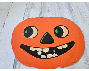 May include: A vintage orange paper pumpkin decoration with a smiling face. The pumpkin has black eyes with green pupils, a black nose, and a toothy grin. The stem is green. A classic Halloween decoration.