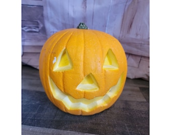 Puede incluir: Una calabaza de Halloween de color naranja brillante con una cara sonriente tallada. La calabaza es de plástico y tiene un tallo verde.
