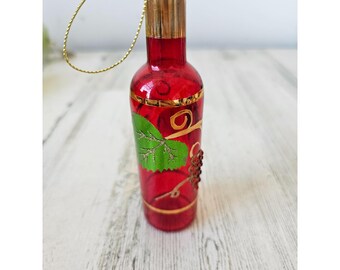 Può includere: Ciondolo a forma di bottiglia di vetro rosso con tappo dorato e una fascia dorata intorno al centro. La bottiglia ha un disegno dipinto in verde e oro di una vite con foglie e uva.