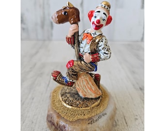 May include: A small, colorful figurine of a clown riding a rocking horse. The clown is wearing a red and white jester hat, a white shirt with a gold vest, and red and white pants. The rocking horse is brown and white. The figurine is sitting on a small, round, beige base with gold accents. The base is signed "Ron 96".