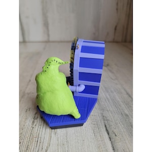May include: A green Christmas ornament featuring the character Oogie Boogie from the movie The Nightmare Before Christmas. Oogie Boogie is sitting in front of a blue and purple chimney.