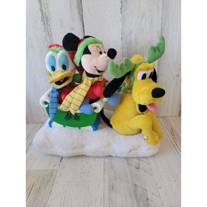 Puede incluir: Un trineo de Navidad de peluche con Mickey Mouse, Donald Duck y Pluto. Mickey lleva un gorro y una bufanda rojos y verdes. Donald lleva una bufanda azul. Pluto lleva un gorro verde con cuernos. El trineo está sobre una superficie blanca y esponjosa.