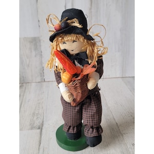 May include: A scarecrow doll with a black hat and brown plaid pants holds a basket of fall harvest vegetables. The doll has a friendly face and is decorated with straw and fall leaves.