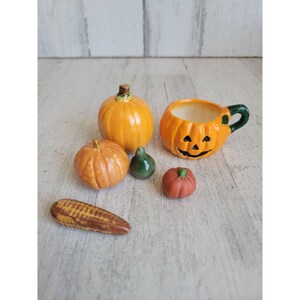 May include: A set of miniature ceramic pumpkins and gourds, including a jack-o'-lantern mug, a small orange pumpkin, a green gourd, and a small orange pumpkin with a stem. The items are arranged on a light wood surface.