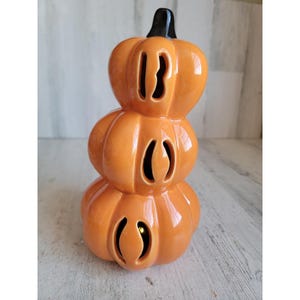 Könnte beinhalten: Gestapelte Keramik-Kürbisdekoration mit glänzendem orangefarbenem Finish. Der Kürbis hat drei Abschnitte mit ausgeschnittenen Augenformen und einen schwarzen Stiel. Im unteren Abschnitt ist ein kleines Licht zu sehen.