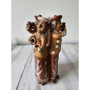 May include: A handcrafted ceramic sculpture depicting three figures. The figures are dressed in brown and tan clothing, with hats and decorative patterns. One figure holds a small bowl. The sculpture is on a white and grey surface.