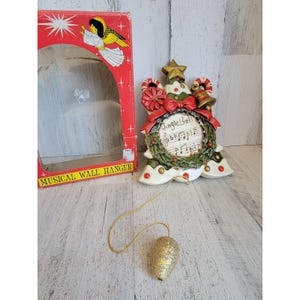May include: A decorative musical wall hanger featuring a Christmas wreath design with a gold star, red bow, and candy canes. The center displays the text Jingle Bell and musical notes. A gold glittery pinecone pull cord hangs from the bottom.