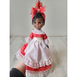May include: A red and white doll with black hair and a red and white polka dot dress. The doll is wearing a white apron with lace trim and a red bow on her head.