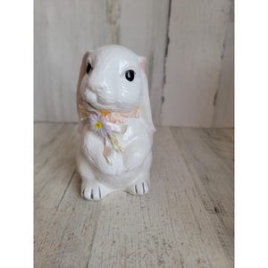 May include: A white ceramic bunny figurine with pink-lined ears and black eyes. A light pink ribbon and a small flower adorn the bunny's neck. The bunny is sitting upright, with its front paws visible. The figurine is on a light-colored wooden surface.