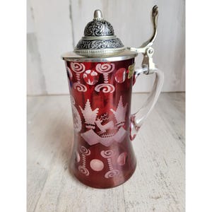 May include: A ruby red glass beer stein with a silver-colored lid and handle. The stein features etched designs of a deer, trees, and decorative patterns. The handle is clear glass.