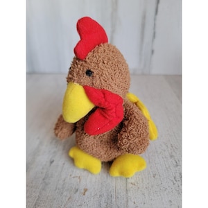 May include: A plush toy rooster with brown, textured fur. It features a red comb and wattle, a yellow beak, and yellow feet. The rooster has small black eyes and a yellow tail feather. The toy is designed to stand upright.