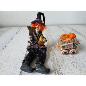 May include: A Halloween witch doll with orange hair, a black hat, and a broom. She holds a carved pumpkin and sits next to a wooden cart filled with pumpkins. The cart has a sign that reads "Pumpkins For Sale."