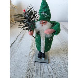 May include: A figurine of a bearded figure wearing a green pointed hat and a matching green coat with a white vertical stripe. The figure has pink hands, black boots, and holds a dark green pine branch with a single red berry on a gray base.