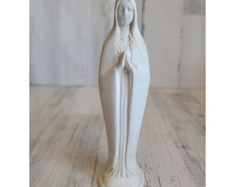 Könnte beinhalten: Weiße bemalte Figur der Jungfrau Maria mit ineinander verschränkten Händen zum Gebet. Die Figur steht auf einer Holzoberfläche.