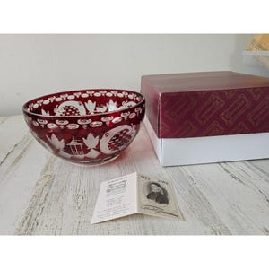 May include: A ruby red glass bowl with intricate white etched designs, including floral and geometric patterns. The bowl is next to a maroon and white box and a small card with text and a portrait. The bowl is a decorative item.