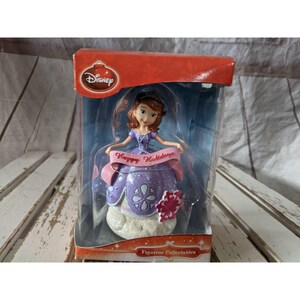 Peut inclure: Une décoration de Noël en forme de princesse Sofia la Première de Disney, en violet et rose. La décoration représente Sofia tenant une bannière sur laquelle est inscrit "Joyeuses Fêtes".